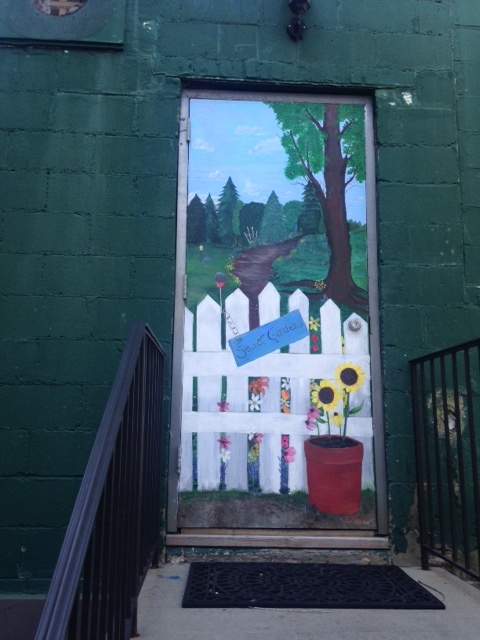 image of a door on which is painted a white picket fence, two sunflowers are in a pot in front of the fence and beyond it is a path that leads to a lovely green meadow